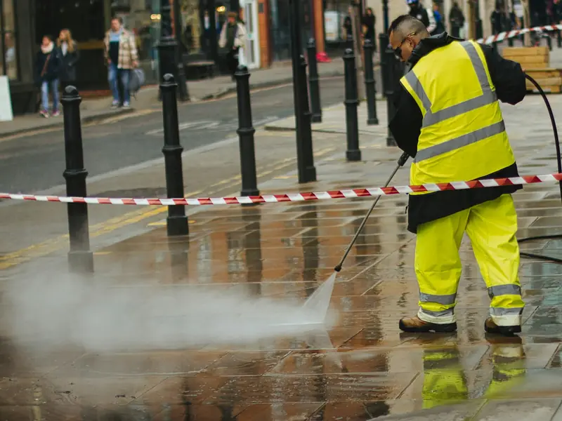 Worker in high-visibility rain gear pressure washing a paved sidewalk behind safety tape