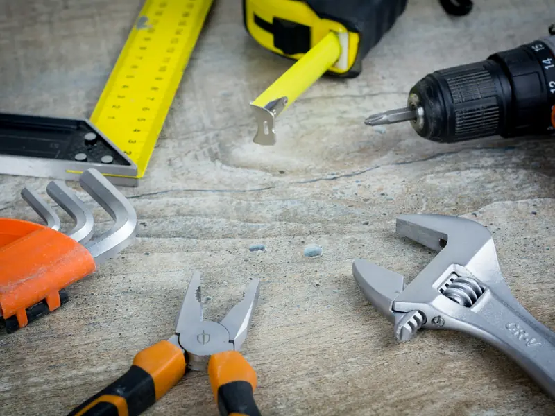 Cordless drill, adjustable wrench, pliers, hex keys, tape measure and framing square laid out on a work surface